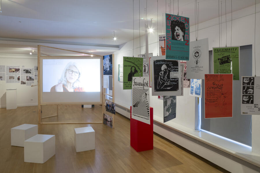 verschiedene Module der Ausstellung: Plakate hänge von der Decke, auf einer Projektionsfläche läuft ein Film und an der Wand hänge Bilder.