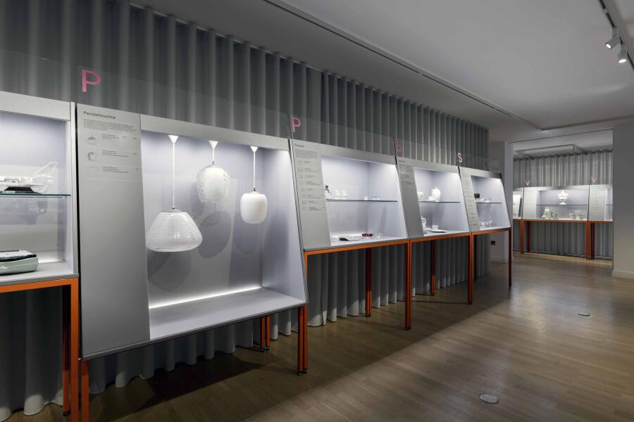 Vor einem hellgrauen Vorhang stehen mehrere graue Vitrinen auf orangenen Beinen. Die Vitrinen sind von innen beleuchtet und zeigen Exponate aus den Sammlungen vom Focke Museum und dem Wilhelm Wagenfeld Haus.