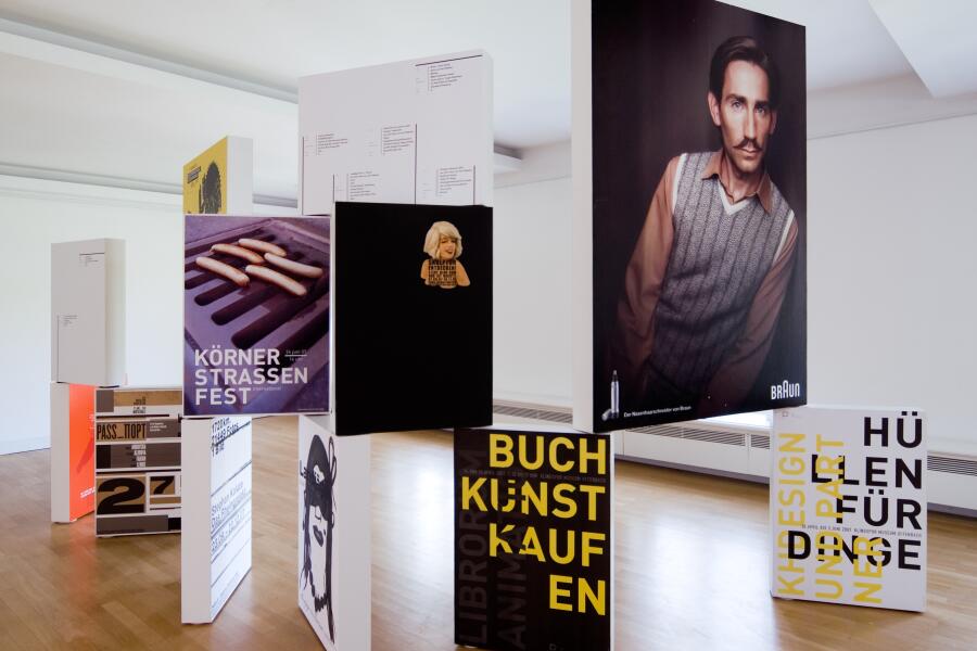 Bunte Plakate wurden auf schmalen, kastenförmigen Bildträgern aufgebracht und stehen nebeneinander und übereinander im Ausstellungraum.