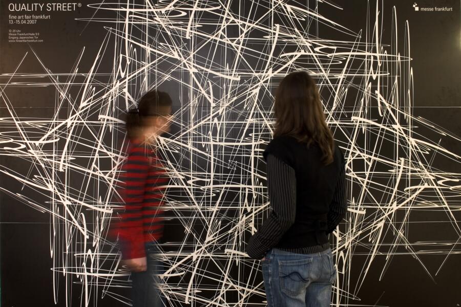 zwei unscharfe Besucherinnen stehen vor einem großformatigen Plakat, das weiße Linien auf schwarzem Grund zeigt.