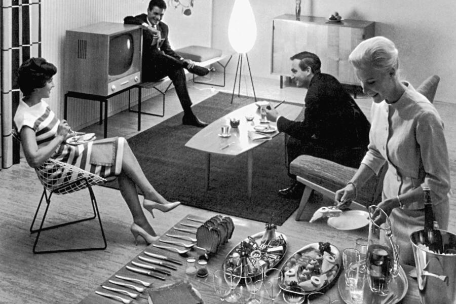 Das schwarz-weiß Foto zeigt drei adrett gekleidete junge Menschen, die einem schicken Wohnzimmer der 1960er Jahre agieren. Auf dem Tisch erkennt man Objekte von Wilhelm Wagenfeld.