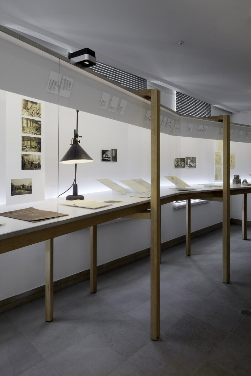 eine diagonal durch den Ausstellungsraum verlaufende Vitrine mit Dokumenten und einer Leuchte